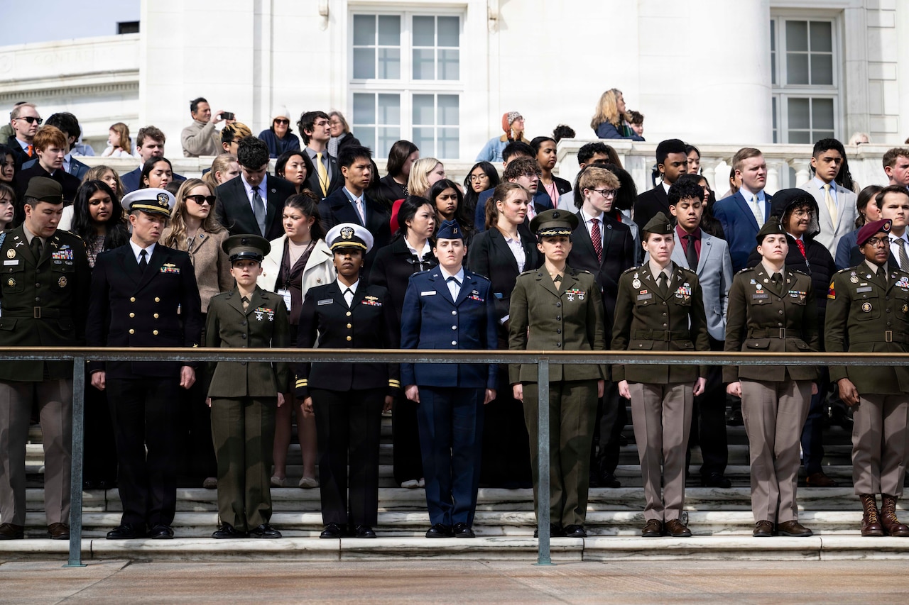 A group of military personnel in dress uniforms stand at attention in front of a group of young adults in business attire as they pose for a photo in front of a building.