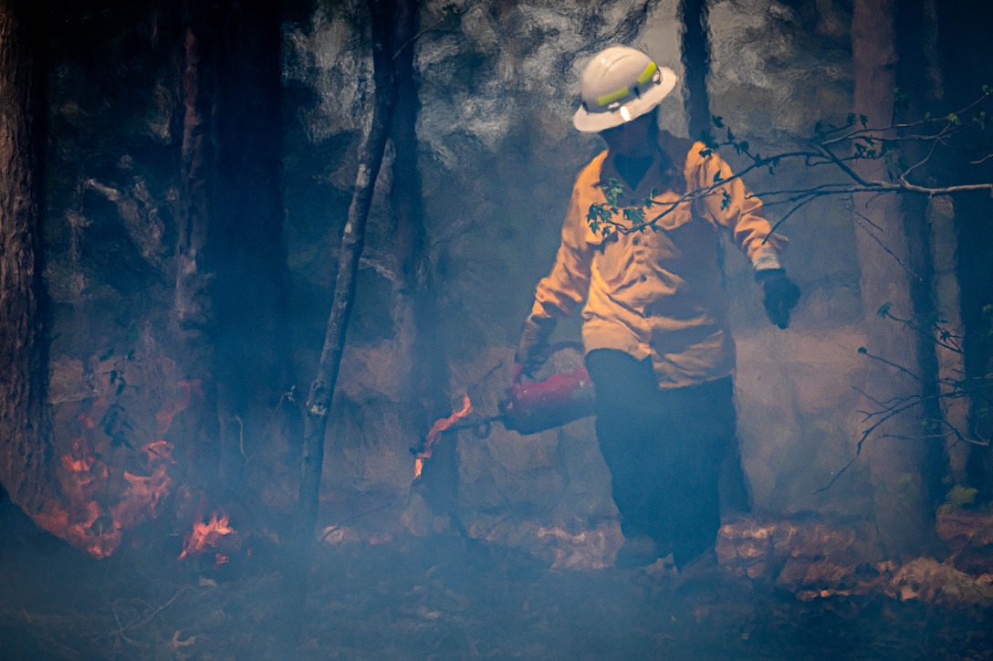 Shows a photo of a women in a yellow shirt and a hard hat holding a gas can with a lit end that is being poured on the ground.