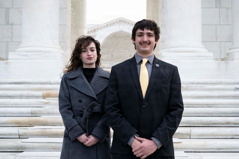A young man and woman in business attire stand on the stairway of a historic building to pose for a photo.