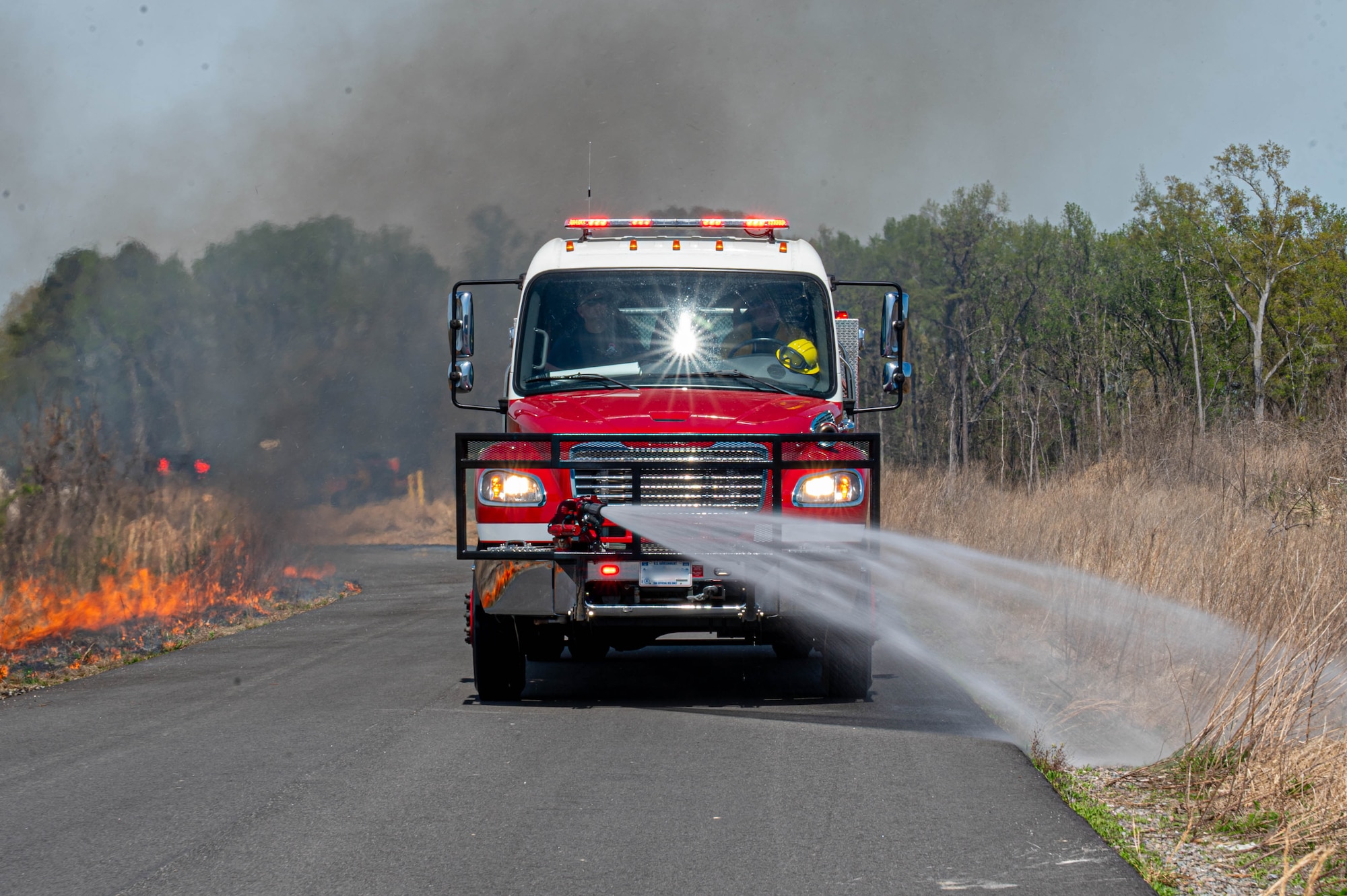 Shows a fire truck spraying water to the right side of the road with a controlled fire on the left side of the road.