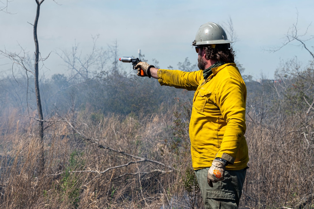 Photo shows a man with a yellow shirt and a gray hard hat holding out a flare gun with one hand holding it out towards brush.
