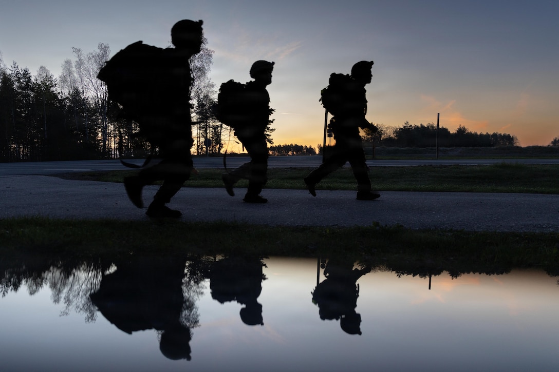 Three soldiers in tactical gear march as the sun rises in the distance as reflected in a puddle in the foreground.