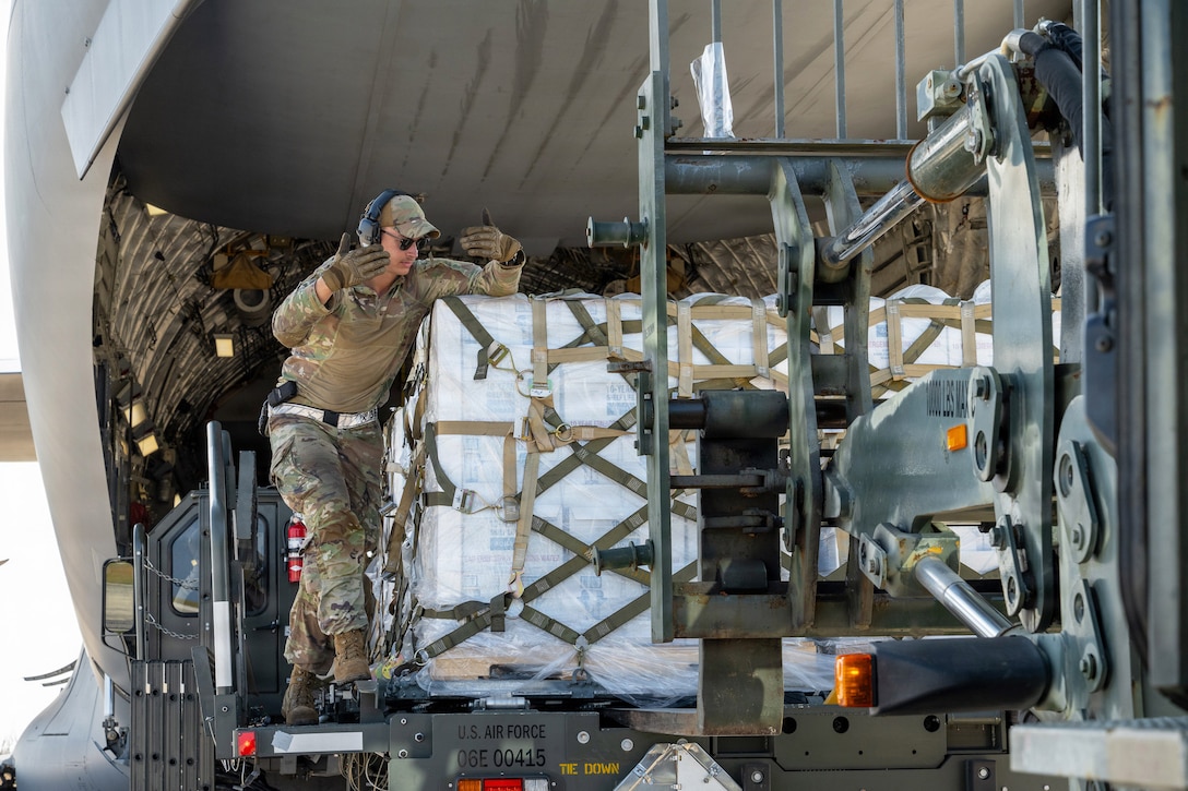 An airman standing next to cargo near an aircraft waves their hands to direct a forklift.