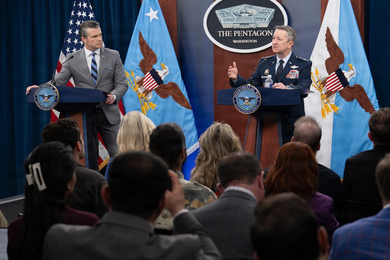 Two men stand behind lecterns; one is wearing a gray suit and looking at the other man wearing a military dress uniform as he speaks to a crowd of people in a room. Behind them are flags against a wall and a sign that reads "The Pentagon — Washington." In the foreground are people in business attire sitting in chairs facing the two men.