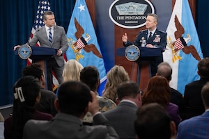 Two men stand behind lecterns; one is wearing a gray suit and looking at the other man wearing a military dress uniform as he speaks to a crowd of people in a room. Behind them are flags against a wall and a sign that reads "The Pentagon — Washington." In the foreground are people in business attire sitting in chairs facing the two men.