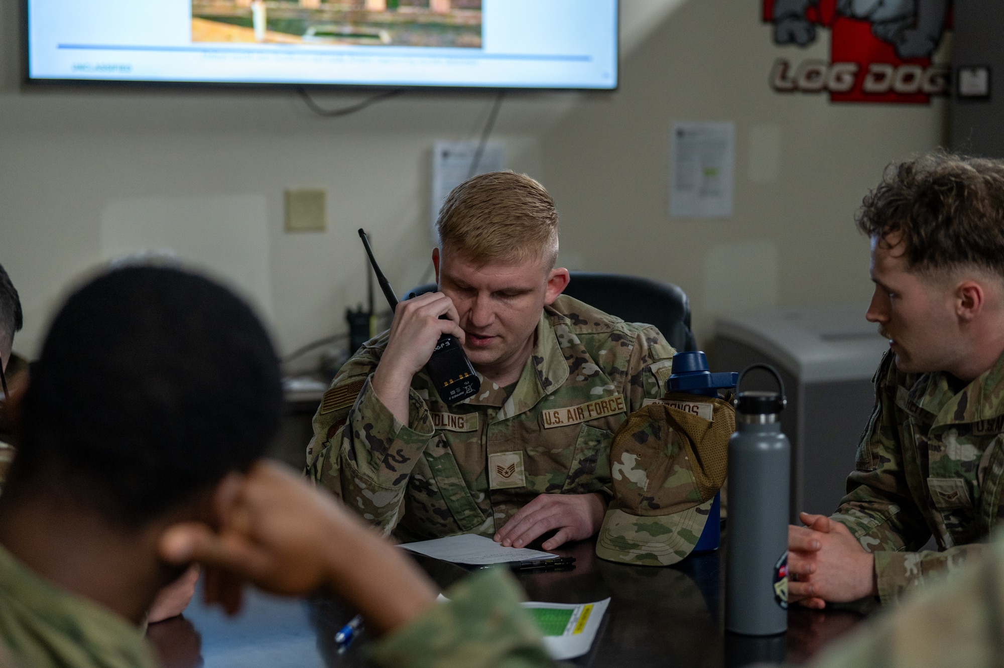 A uniformed service member speaks into a handheld radio while sitting among a group during a training event.