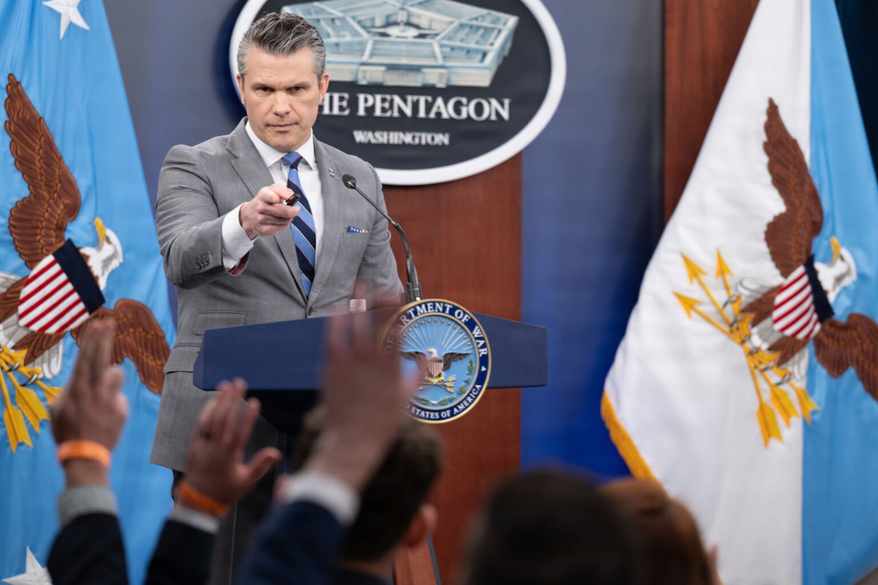 A man stands behind a lectern and points out at other people seated before him, who have their hands raised. Behind him is a sign that reads "The Pentagon — Washington."