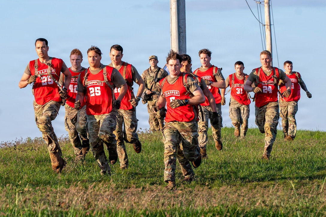 A group of competitors wearing red jerseys over their uniforms run in a field under a blue sky.