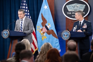 Two people, one in a business suit and the other in a dress military uniform, speak at lecterns to a seated audience.