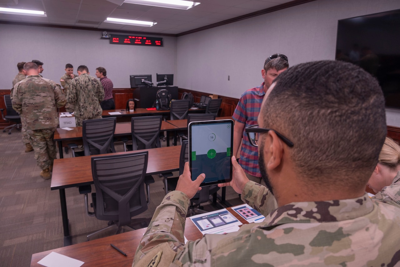 A man in a camouflage military uniform holds a tablet in front of himself while participating in a medical study in a classroom; there are several other people in similar attire and casual attire standing in the room.