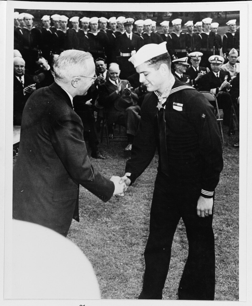 Two men, one in a suit and one in a military dress uniform wearing a medal, shake hands as people behind them, seated in chairs, clap.