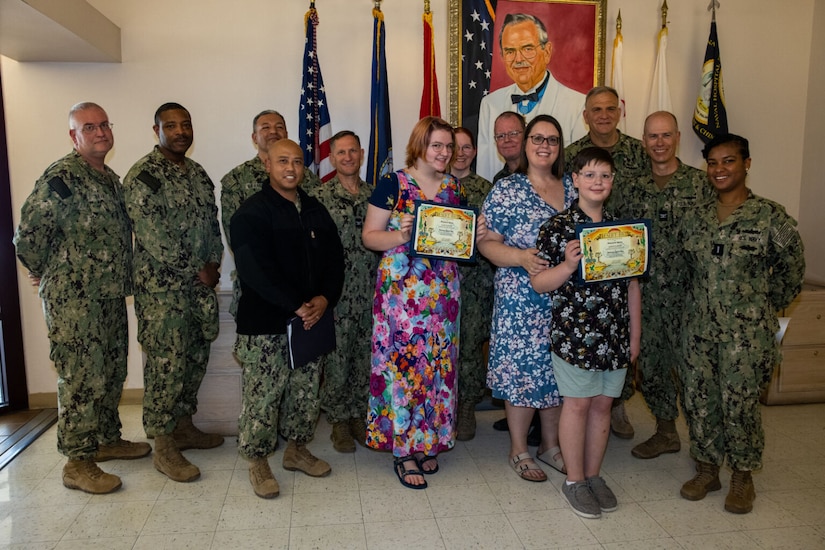 Two women and a boy in casual attire pose for a photo at a hospital with people in camouflage military uniforms as they stand in front of flags and a painting of a man wearing a blue medal; the boy and one of the women are holding certificates.