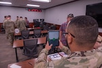A man in a camouflage military uniform holds a tablet in front of himself while participating in a medical study in a classroom; there are several other people in similar attire and casual attire standing in the room.