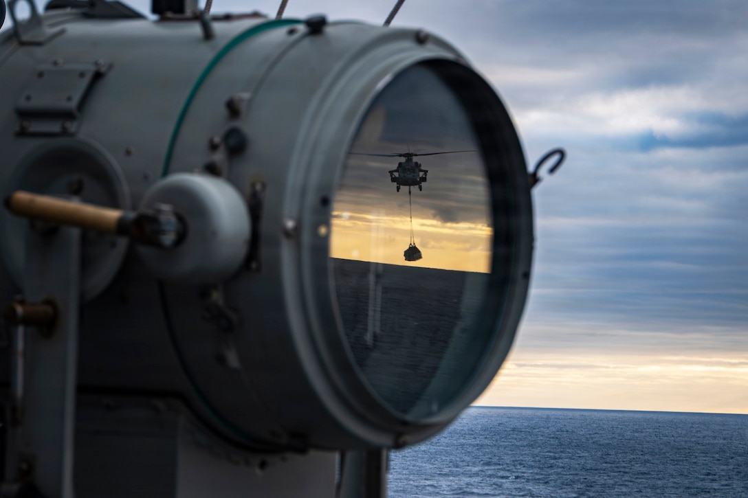 A helicopter carries cargo while flying above a body of water in a sunlit sky as seen through a ship's searchlight.