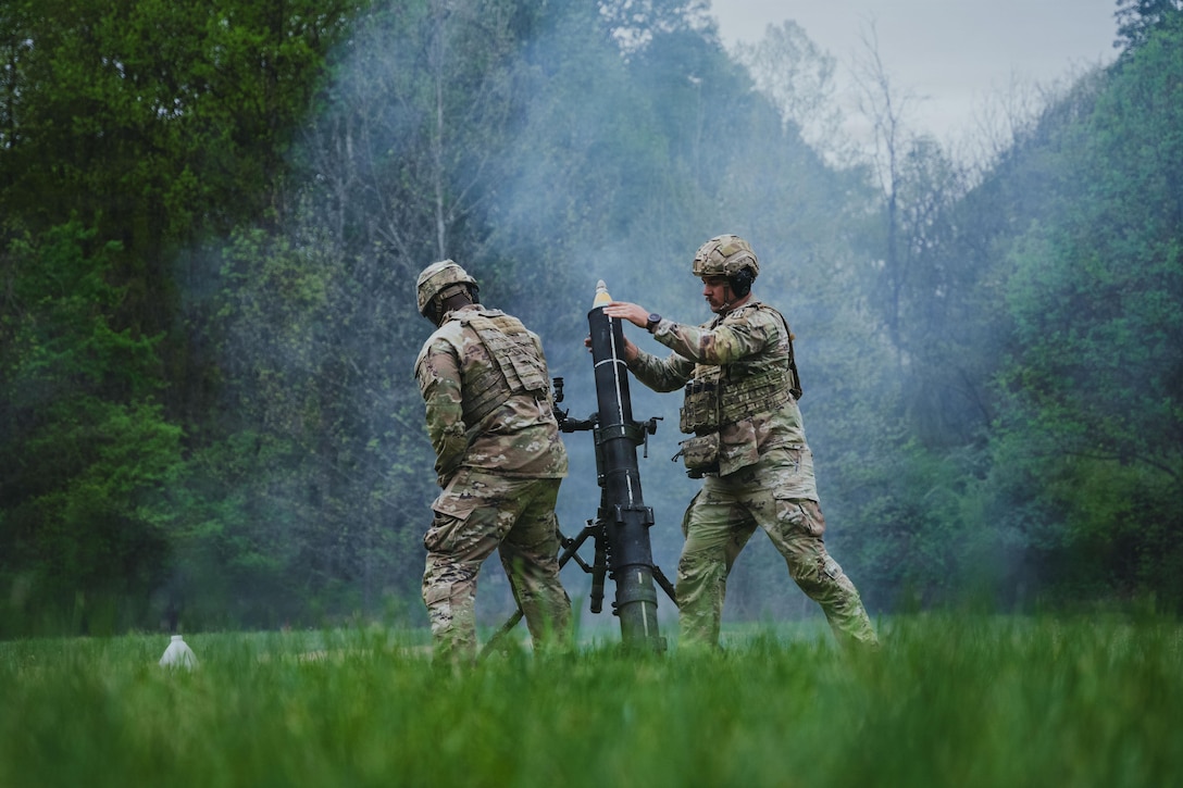 Two soldiers stand in a grassy area with a mortar system, with smoke in the air and trees in the background.