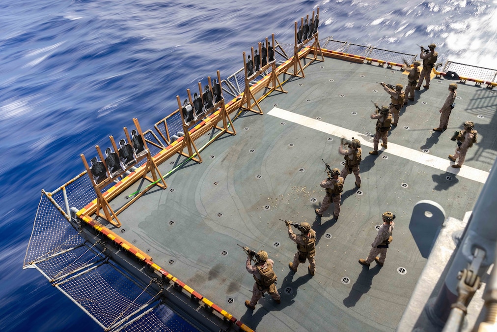 Service members standing in a row aboard a ship point guns at targets used for training.