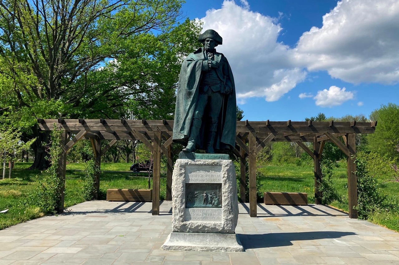 A greenish sculpture depicts a man in uniform on a pedestal in a park-like setting.