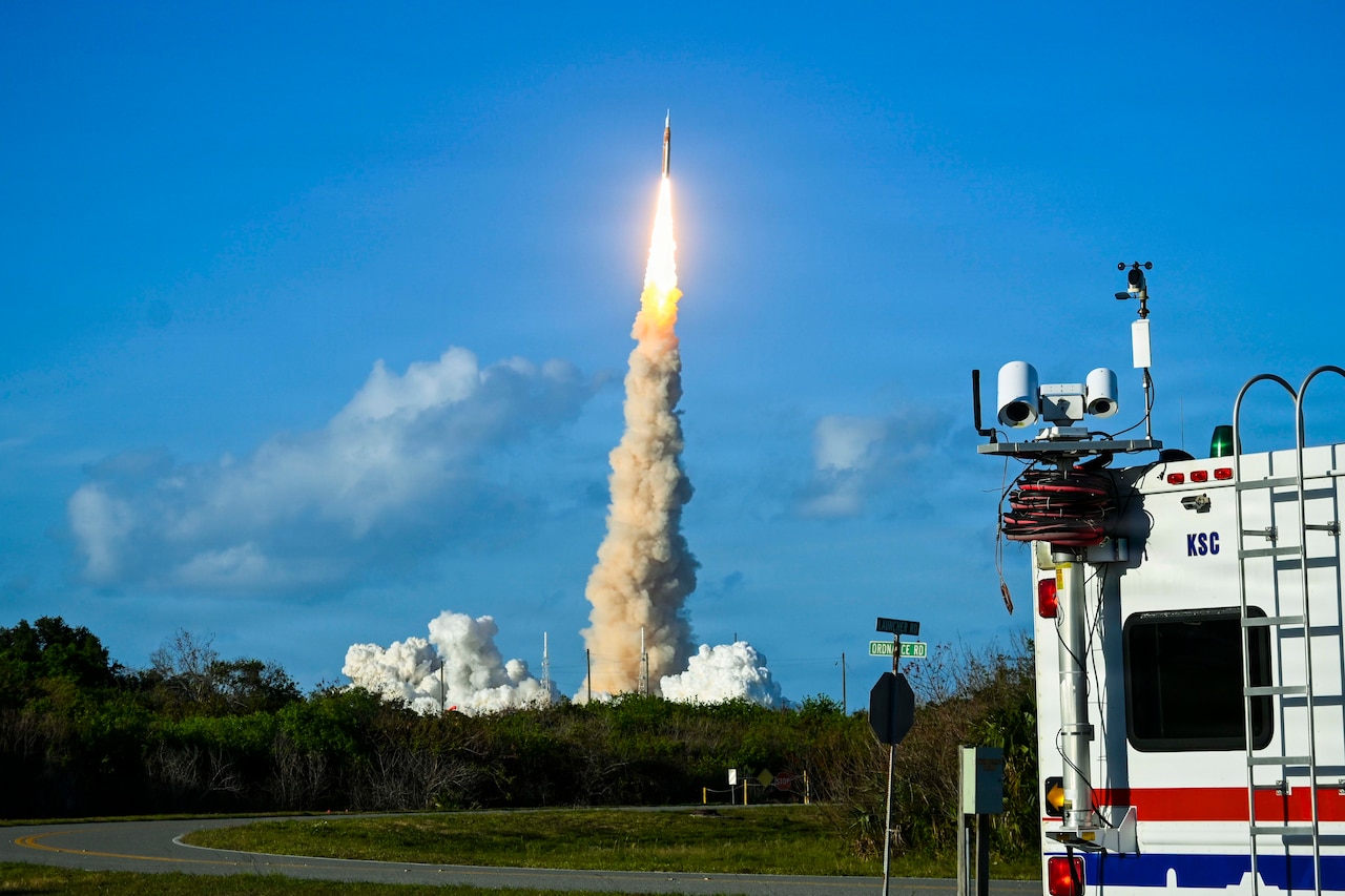 A rocket launches leaving behind a plume of smoke against a partly cloudy sky in the distance and an emergency medical vehicle in the foreground.