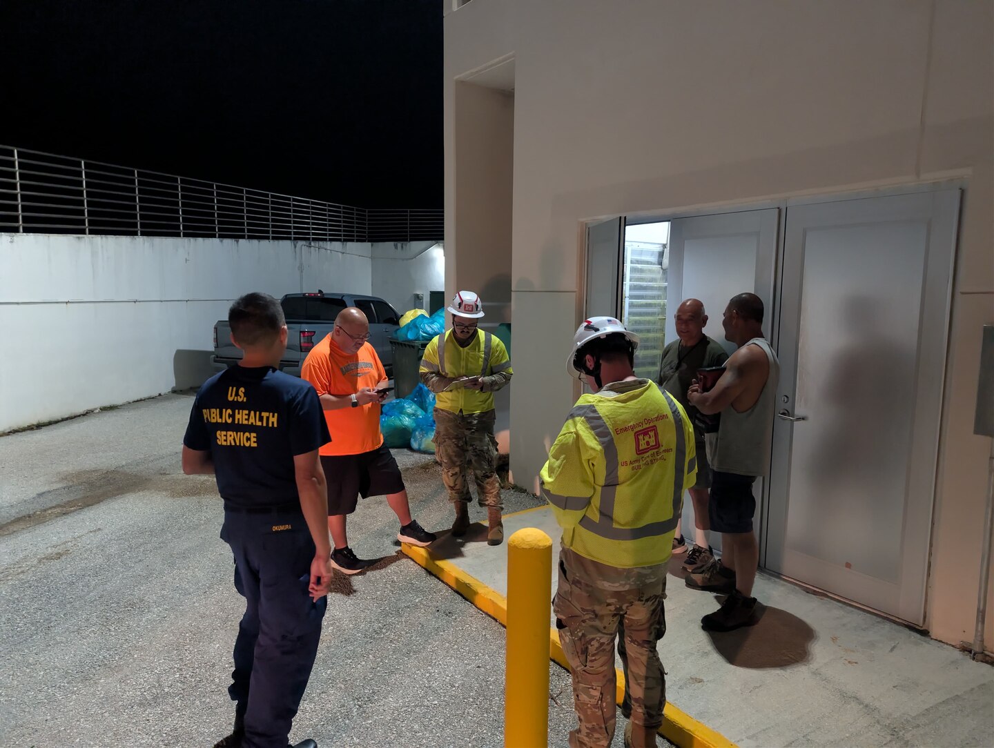two soldiers are with a group of people outside an entrance looking at their phones.