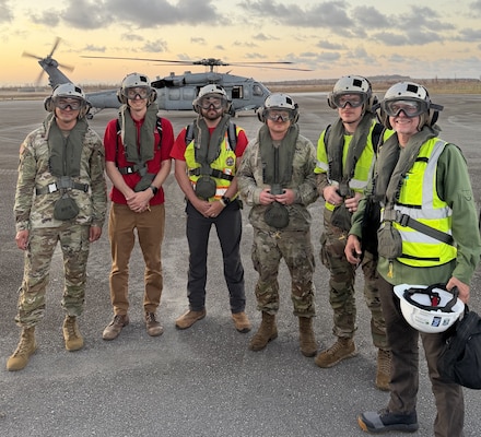 A group of Soldier and civilians wearing flight gear stand in front of a military helicopter.