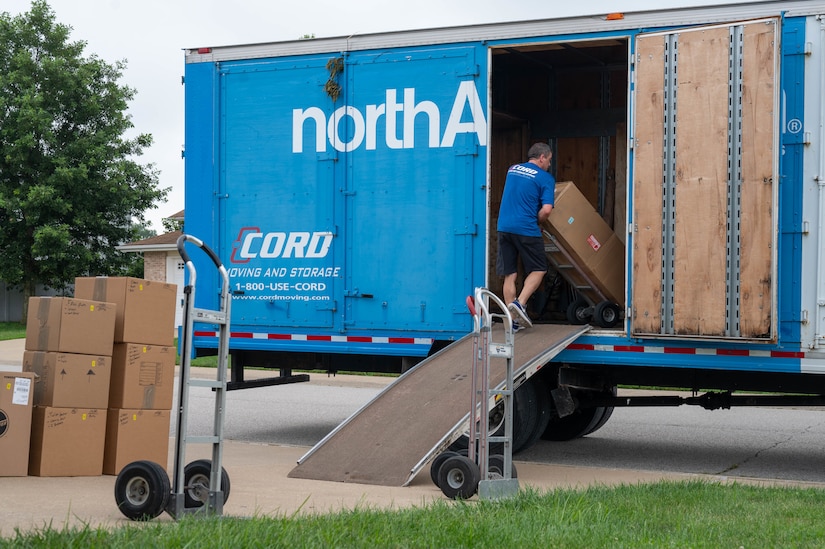 A man loads boxes on a blue moving truck; stacks of boxes are on a driveway next to the truck.