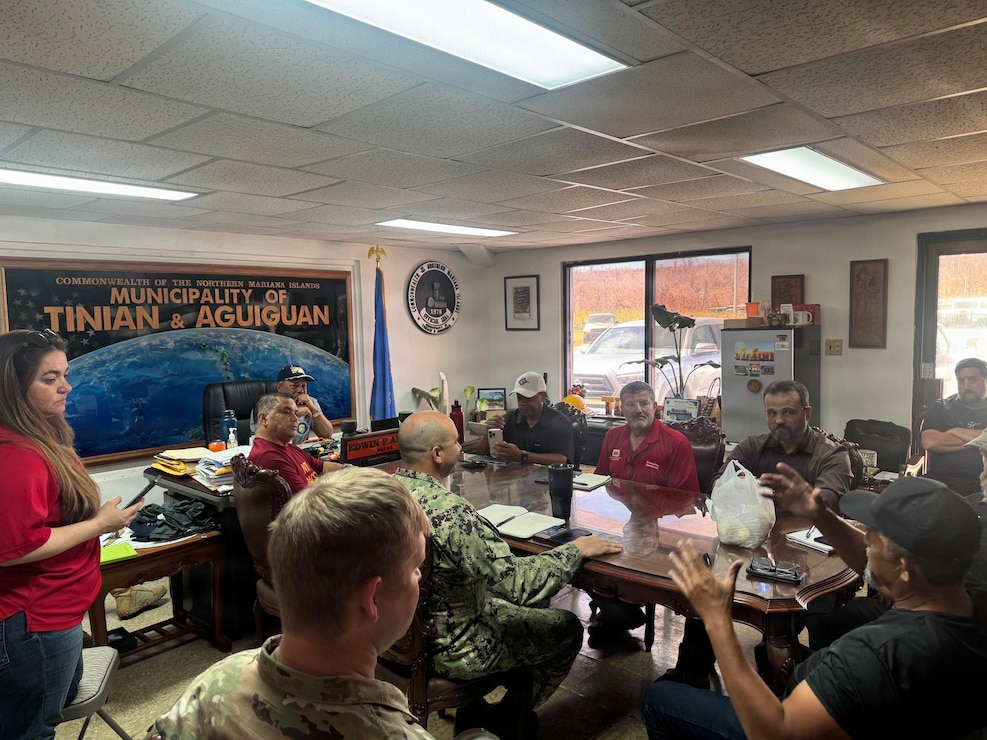 A group of individuals sit around a large wooden conference room table. On the wall a large mural of the pacific ocean hangs with the words Municipality of Tinian & Aguiguan written on it.