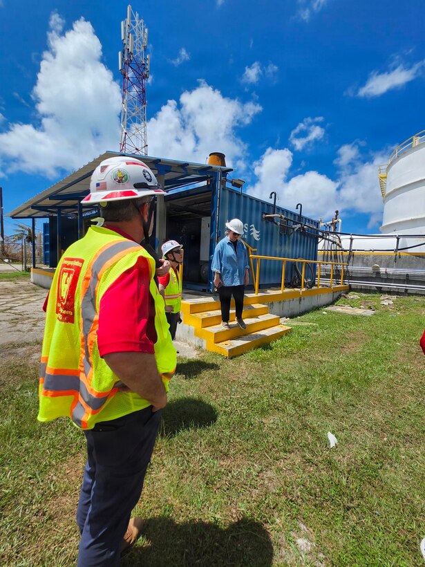 A man in a hard hat and safety vest looks at a person coming down stairs at a large fuel facility