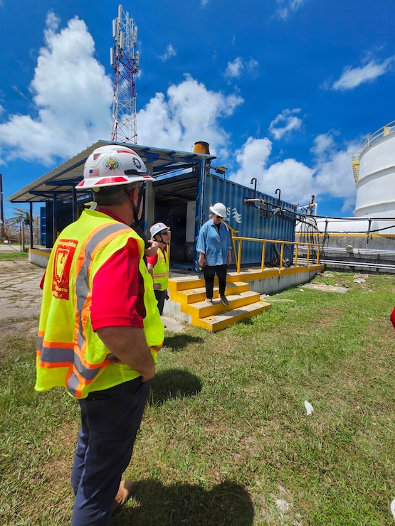 A man in a hard hat and safety vest looks at a person coming down stairs at a large fuel facility