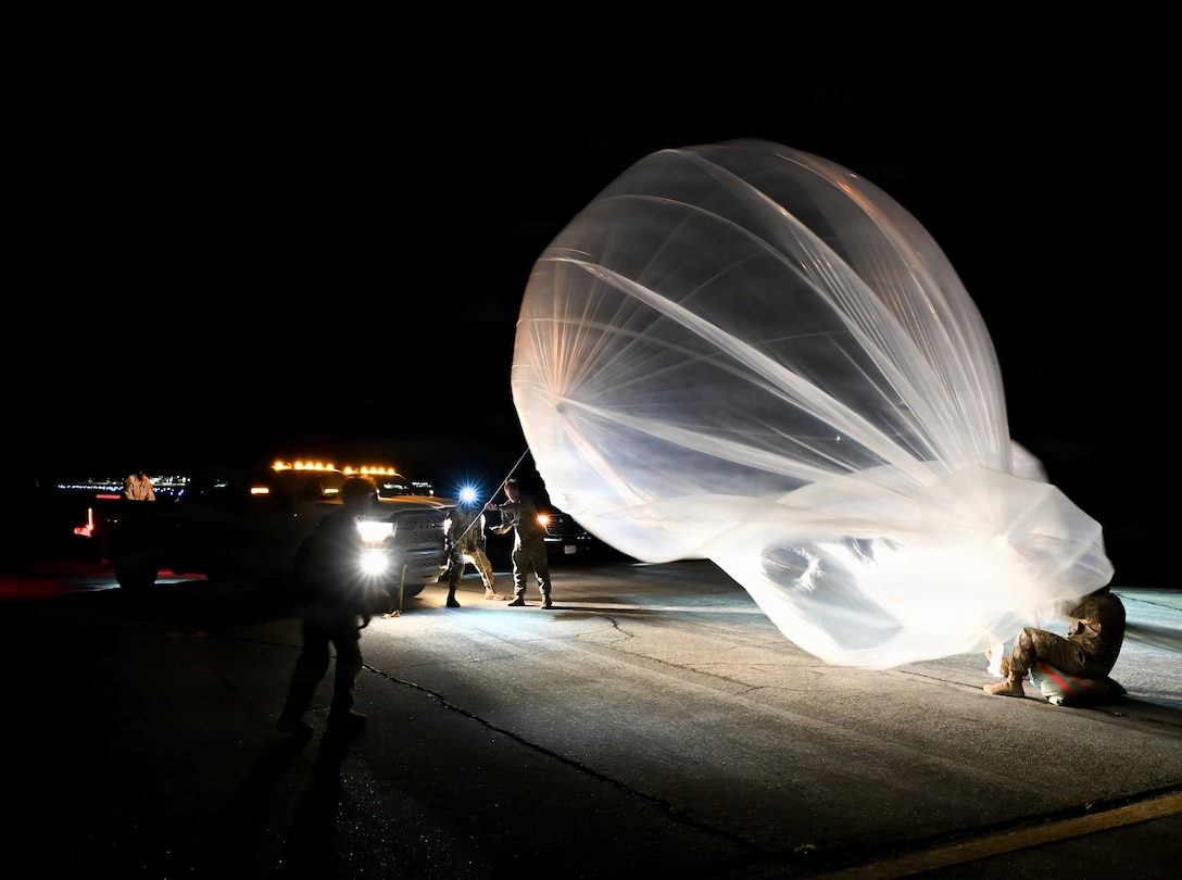 Soldiers prepare to release a white balloon into a dark sky while standing in front of pickup trucks shining their headlines.
