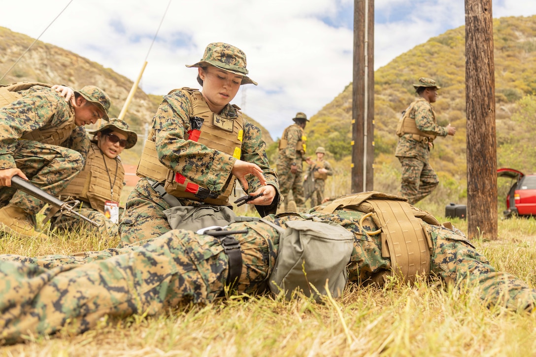 A sailor works on a dummy lying in a field as fellow sailors train in the background during the day.