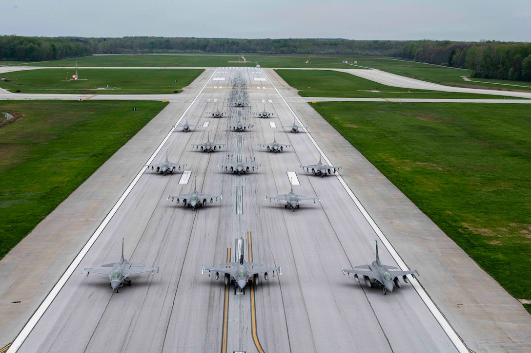Sixteen aircraft move in formation on a tarmac with a wooded area in the background on a gloomy day.