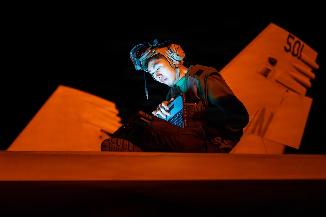 A sailor sits on an aircraft in the dark while using a laptop.