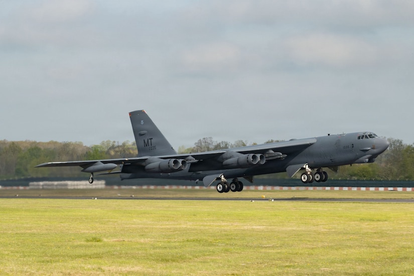 A large military bomber aircraft takes off from a runway into a cloudy sky with trees in the background.