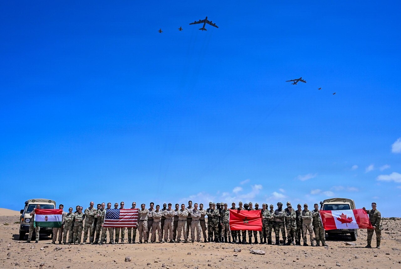 Dozens of people in camouflage military uniforms pose for a photo in the desert while holding up four flags as six large military aircraft fly overhead.