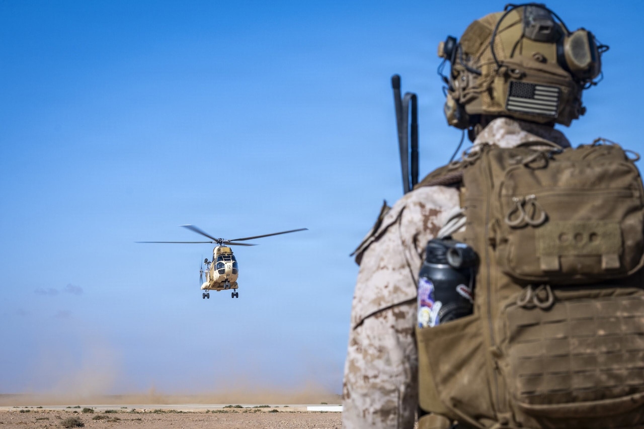 A person in a camouflage military uniform with an American flag patch on their helmet watches a military helicopter as it flies in the air over a desert.