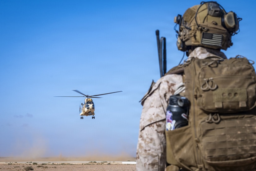 A person in a camouflage military uniform with an American flag patch on their helmet watches a military helicopter as it flies in the air over a desert.