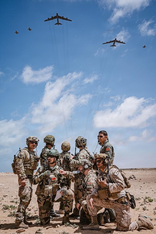 Eight men in camouflage military uniforms, helmets and sunglasses and another man in a flight suit, pose for a photo in a desert location, as five large military aircraft fly overhead.