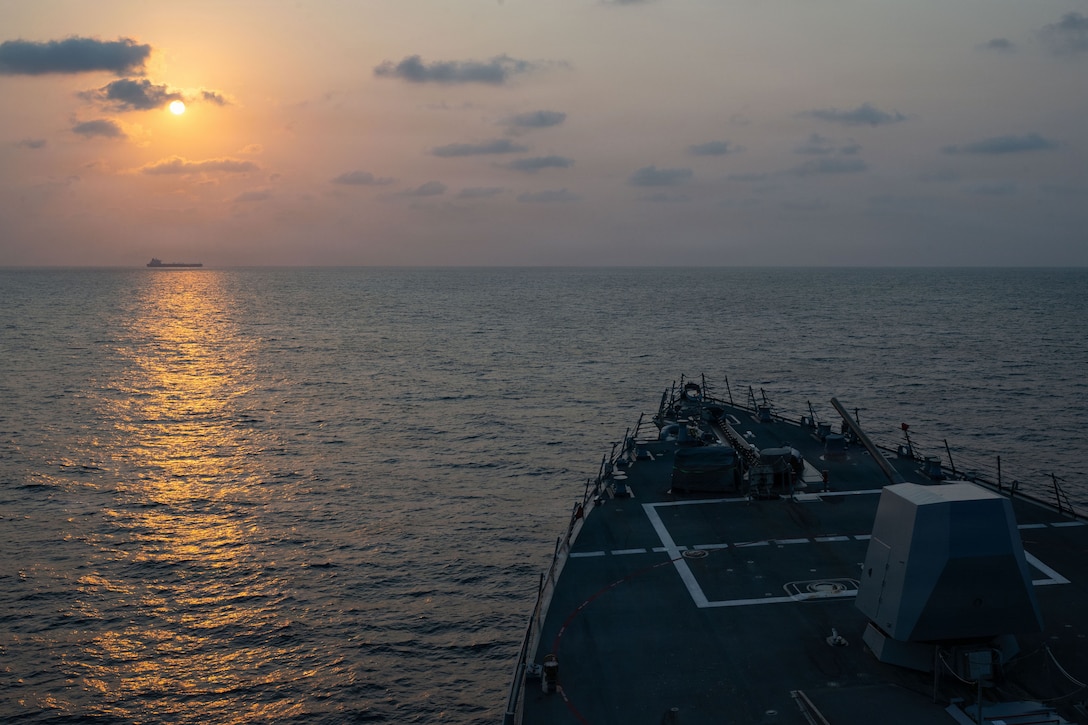 The front of a ship is visible as it operates in water against an orangish sky, with another ship in the distance.