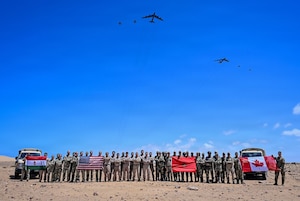 Dozens of people in camouflage military uniforms pose for a photo in the desert while holding up four flags as six large military aircraft fly overhead.