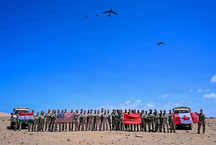 Dozens of people in camouflage military uniforms pose for a photo in the desert while holding up four flags as six large military aircraft fly overhead.