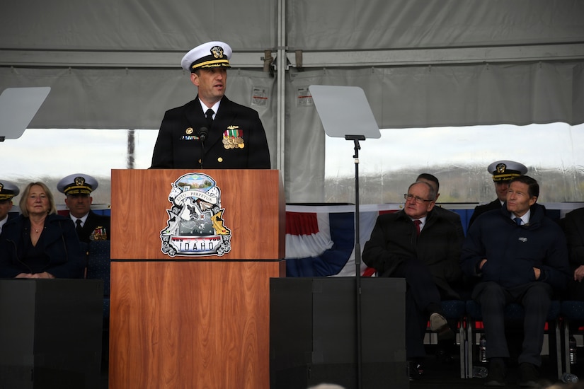 A man in military dress uniform stands at a lectern addressing a crowd of people while people in similar attire and business attire are seated behind him.
