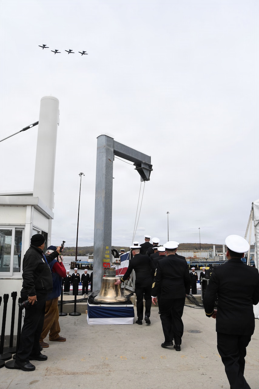 People dressed in military dress uniforms stand in line to ring a large golden bell as military aircraft fly overhead.