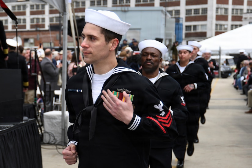 A group of men dressed in military dress uniforms walk in a line between a crowd of people.
