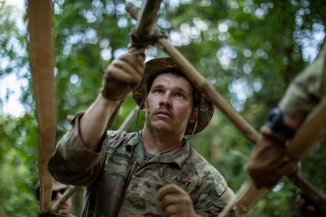 A soldier secures pieces of bamboo together outdoors, with trees in the background.