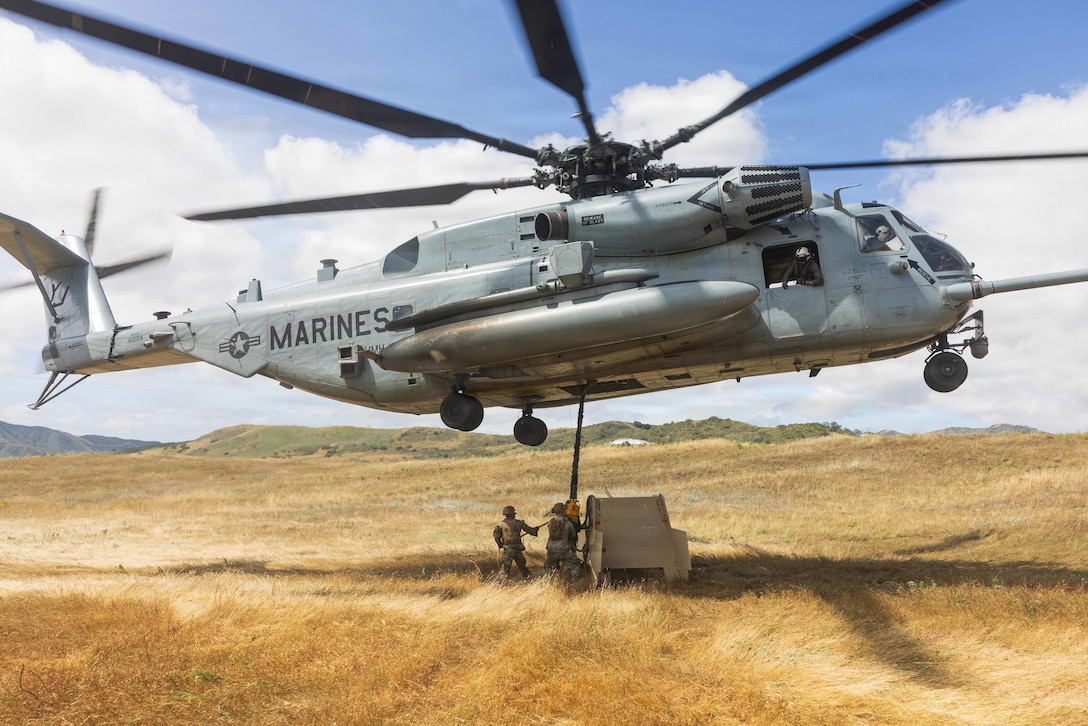 Two Marines attach a steel target to a helicopter hovering above them in a field of golden grass.