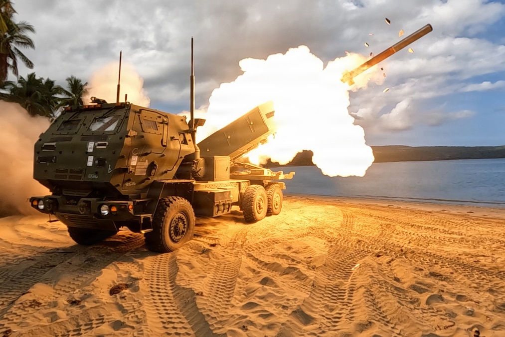 A rocket is launched from a military vehicle on a beach, with palm trees in the background.