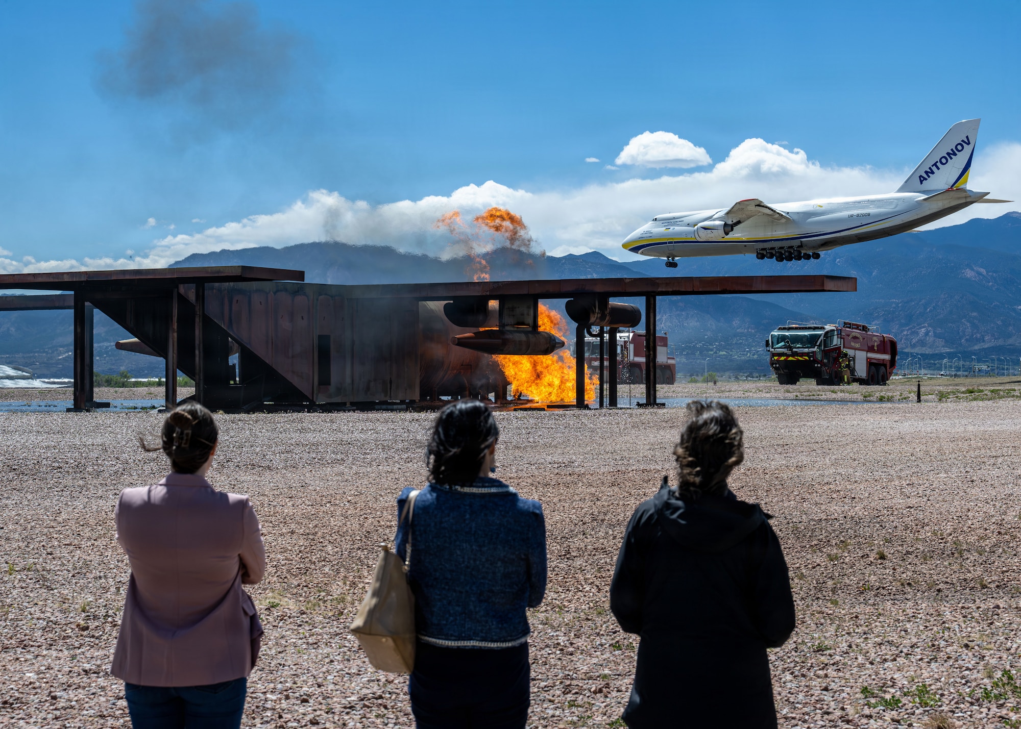 Members of a tour group watch firefighters put out a training fire while a plane flies overhead.