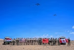 Dozens of people in camouflage military uniforms pose for a photo in the desert while holding up four flags as six large military aircraft fly overhead.