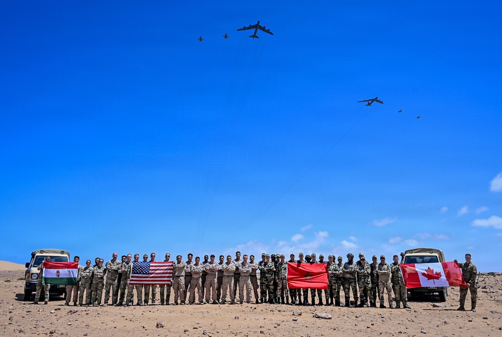 Dozens of people in camouflage military uniforms pose for a photo in the desert while holding up four flags as six large military aircraft fly overhead.