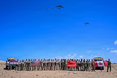 Dozens of people in camouflage military uniforms pose for a photo in the desert while holding up four flags as six large military aircraft fly overhead.